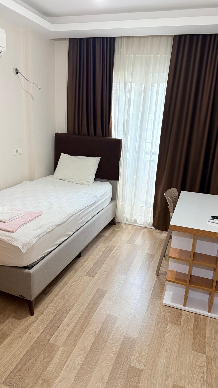 Un lit simple avec une tête de lit, une petite table avec une chaise et une fenêtre avec des rideaux.