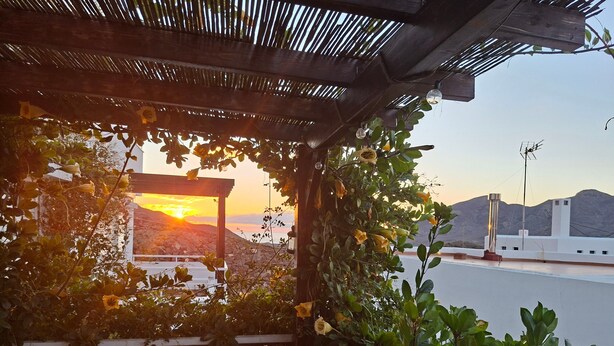 Een overdekt buitengebied met uitzicht op de zonsondergang, een pergola en een wit gebouw op de achtergrond.
