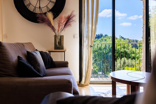 A living room with a sofa, a small table, a clock on the wall, and a view of greenery through a balcony door.