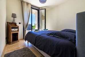 A bedroom with a bed, a nightstand, a lamp, and a view of the outdoors.