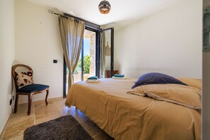 A bedroom with a bed, a chair, a window with curtains, and a view of greenery outside.
