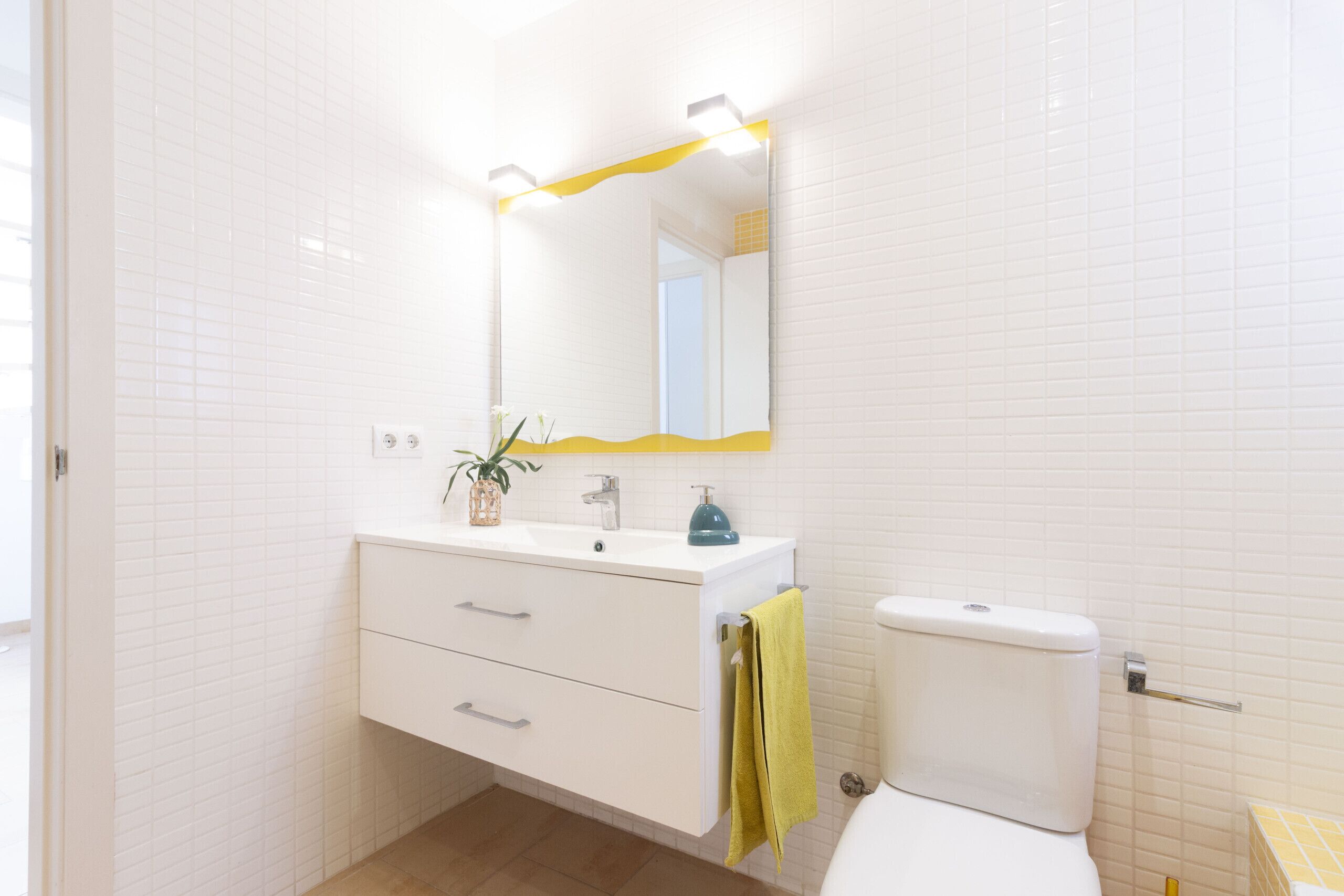 Een badkamer met witte betegelde wanden, een wit wastafelmeubel met spiegel erboven, een gele handdoek aan de muur, en een wit toilet.