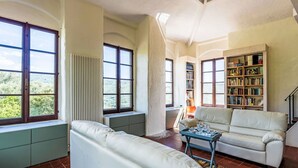 Un salon spacieux avec un canapé en cuir blanc, une table basse bleue et une bibliothèque remplie de livres. De grandes fenêtres offrent une vue sur l’extérieur.