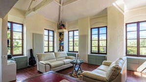 Un salon spacieux avec un canapé beige, une table basse et une bibliothèque. De grandes fenêtres laissent entrer la lumière naturelle et offrent une vue sur la verdure.