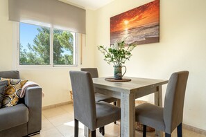 A dining area with a wooden table, grey upholstered chairs, a vase with flowers, and a sunset painting on the wall.