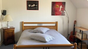 A bedroom with a wooden bed, two pillows, a nightstand with a lamp, a guitar, and two framed pictures on the wall. - ROOM RENTAL for events on the Sarthe racetrack (Change)
