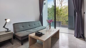 Un salon moderne avec un canapé, une table basse en bois, un bureau avec une lampe et une vue sur la verdure grâce à de grandes fenêtres.