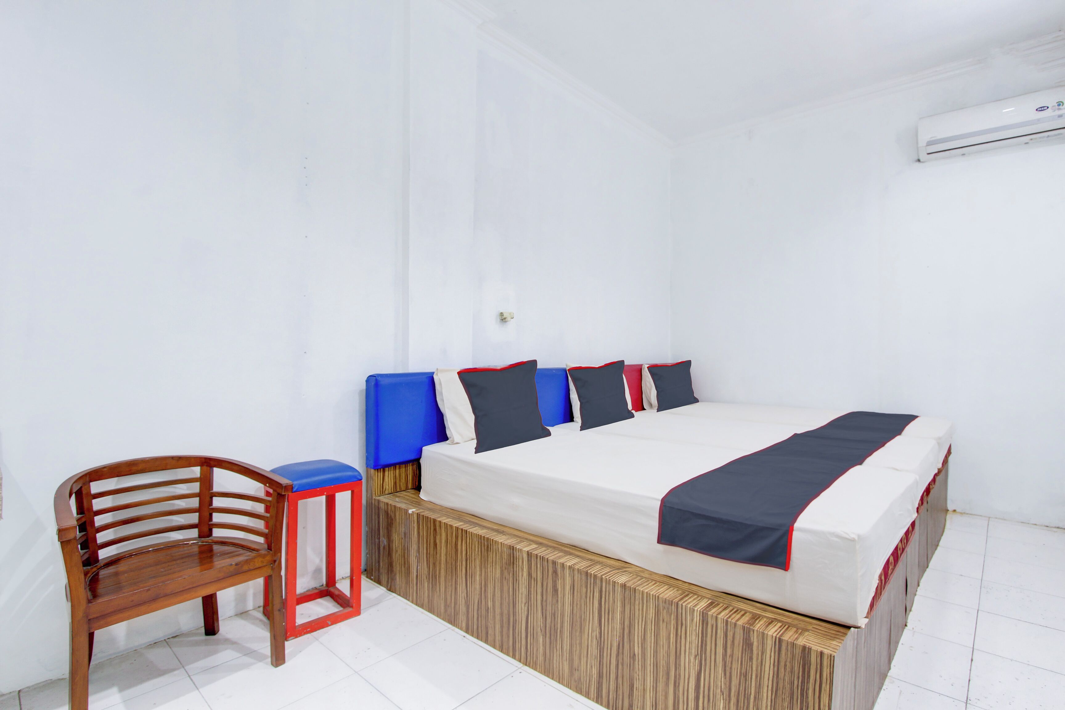 Un dormitorio con estructura de cama de madera, una silla de madera sencilla y un taburete pequeño rojo y azul.