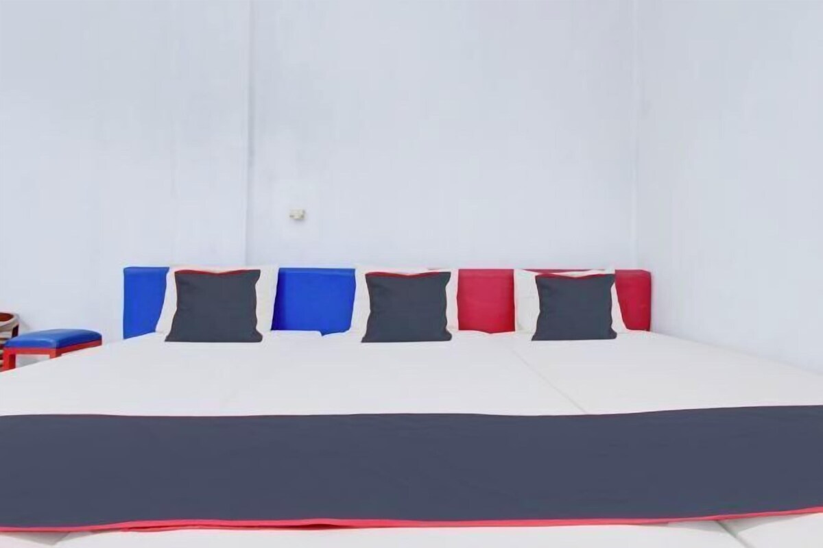 Una cama con colchón blanco y colcha azul oscuro. Hay seis almohadas en colores alternados de azul, rojo y gris.
