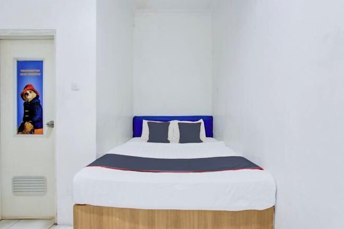 Una cama pequeña y ordenada, con cabecera azul y ropa de cama blanca, un póster en la puerta y una pared blanca.