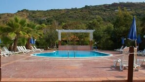 Piscina com pergolado, rodeada por espreguiçadeiras e guarda-sóis, com vista para uma encosta.