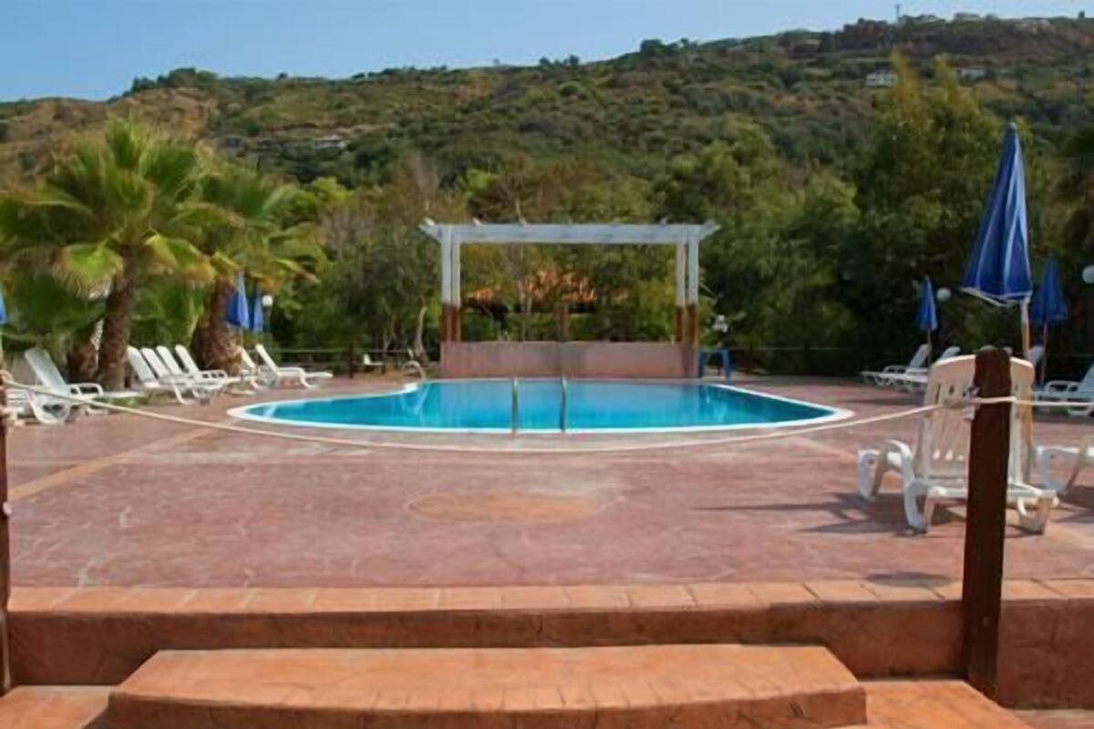 A swimming pool with a pergola, surrounded by lounge chairs and umbrellas, set against a hillside backdrop.