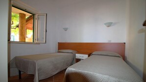 Quarto com duas camas de solteiro, cabeceira de madeira, pequena mesa de cabeceira e janela com vista para área verde.
