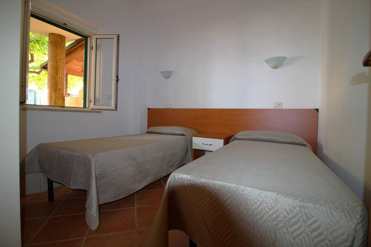A room with two single beds, a wooden headboard, a small bedside table, and a window with a view of greenery.