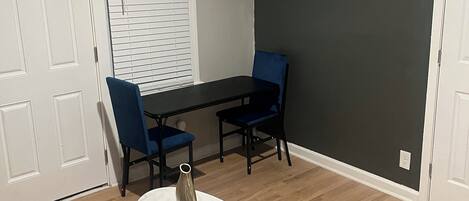 A room with a dining table, two chairs, a small rug, a vase, and a TV mounted on the wall.