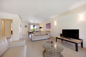 Una sala de estar moderna con un sofá, una mesa de centro de cristal, un televisor sobre un mueble de madera y un comedor al fondo.