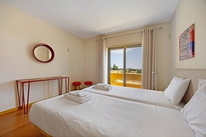 Une chambre d’hôtel avec deux lits, une petite table, deux chaises rouges, un miroir rond et une grande fenêtre avec des rideaux.
