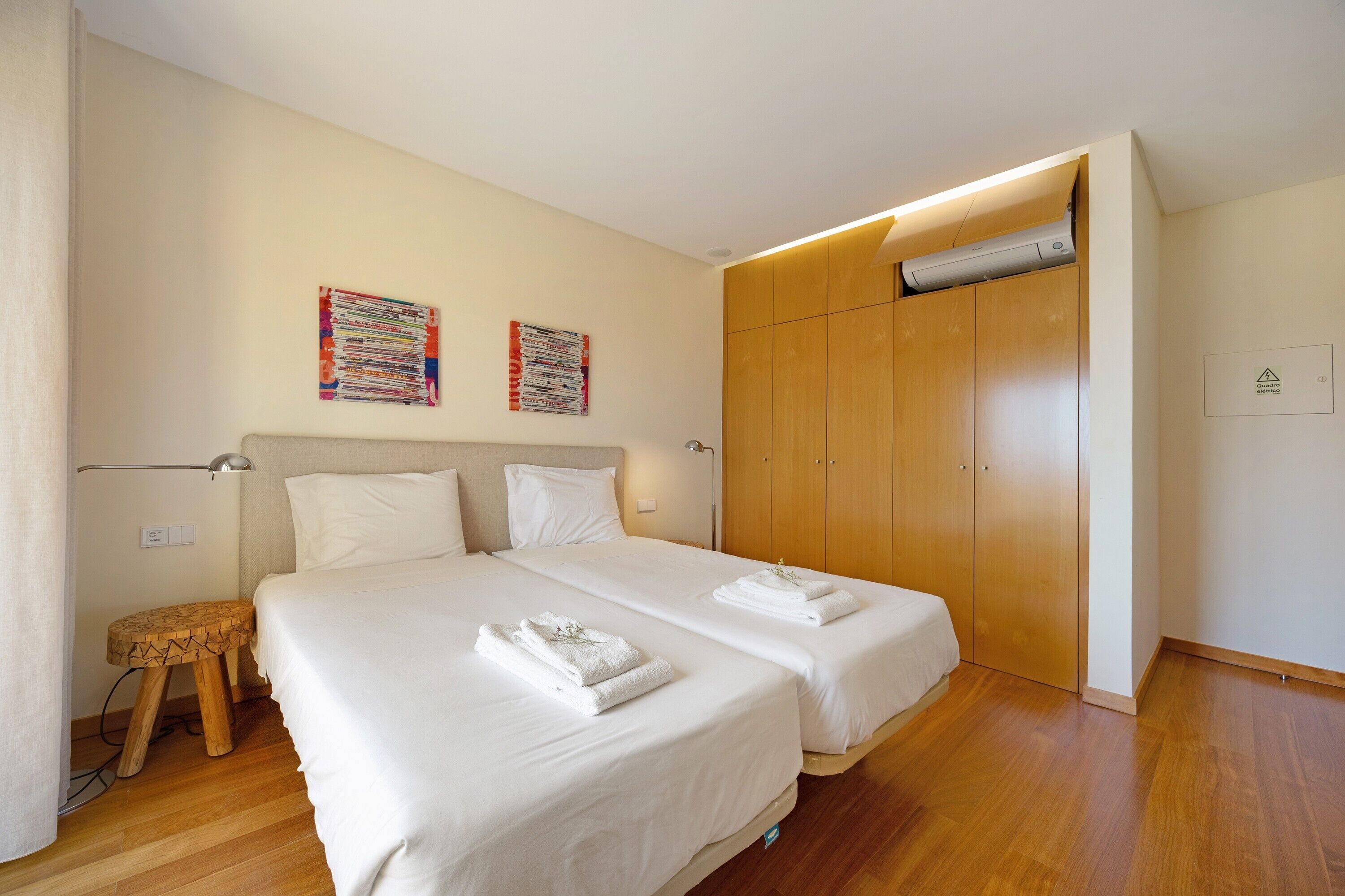 Una camera d'albergo con due letti, un armadio in legno, un piccolo sgabello e due opere d'arte appese al muro.