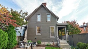 Een tweelaags huis met een overdekte veranda, een pad van stenen en een klein terras met tuinmeubilair.