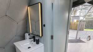 Ein Badezimmer mit weißem Waschbecken, einem Spiegel mit Beleuchtung und einer Tür zu einem Außenbereich.