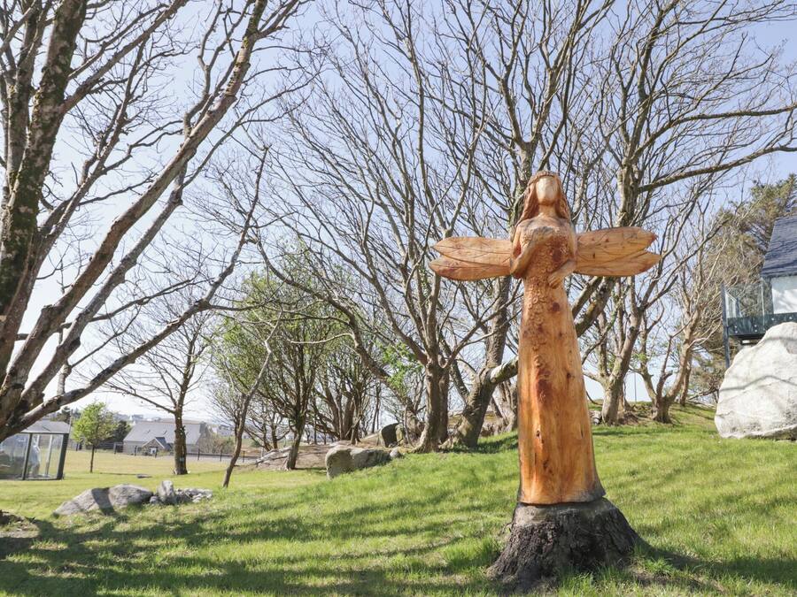 A wooden sculpture of a woman with outstretched arms in a park with trees and rocks.