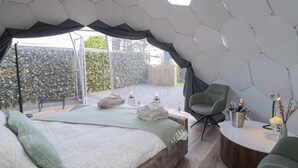 Ein modernes Schlafzimmer mit sechseckiger Decke, einem großen Bett mit Kissen, einer Sitzecke mit einem Sessel und einem kleinen Tisch mit einer Vase und einer Flasche.