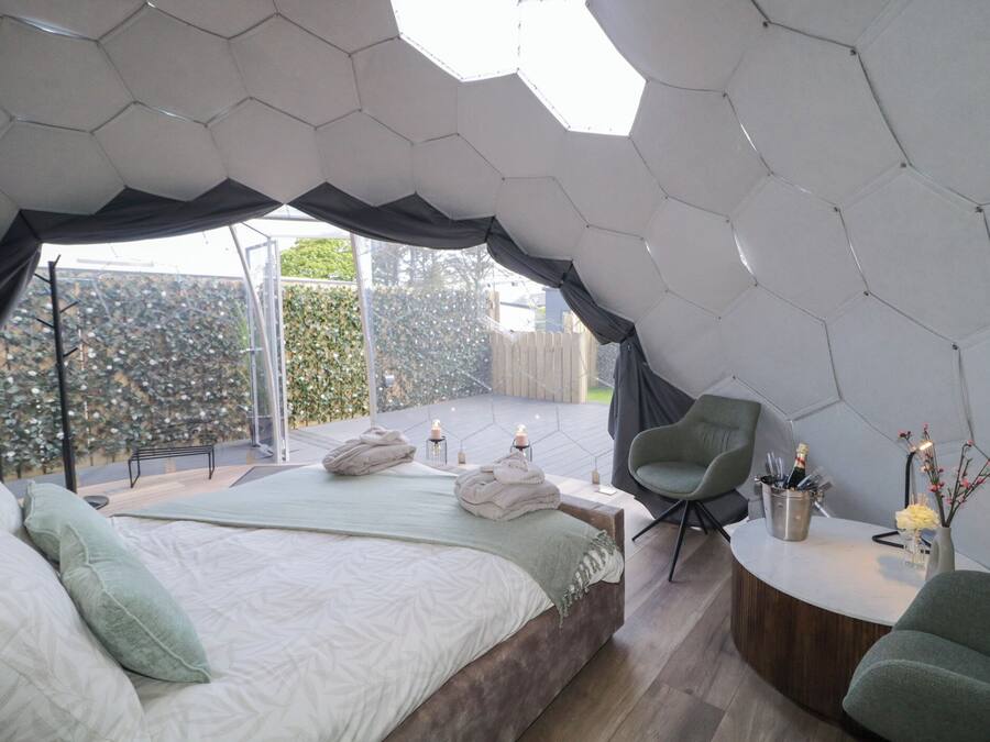 Un dormitorio moderno con techo hexagonal, una cama grande con almohadas, una zona de estar con una silla y una mesita con un florero y una botella.