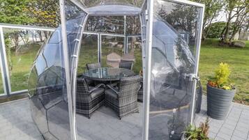 En öppen paviljong med glasbord och flätade stolar, omgiven av en stenlagd yta och krukväxter.