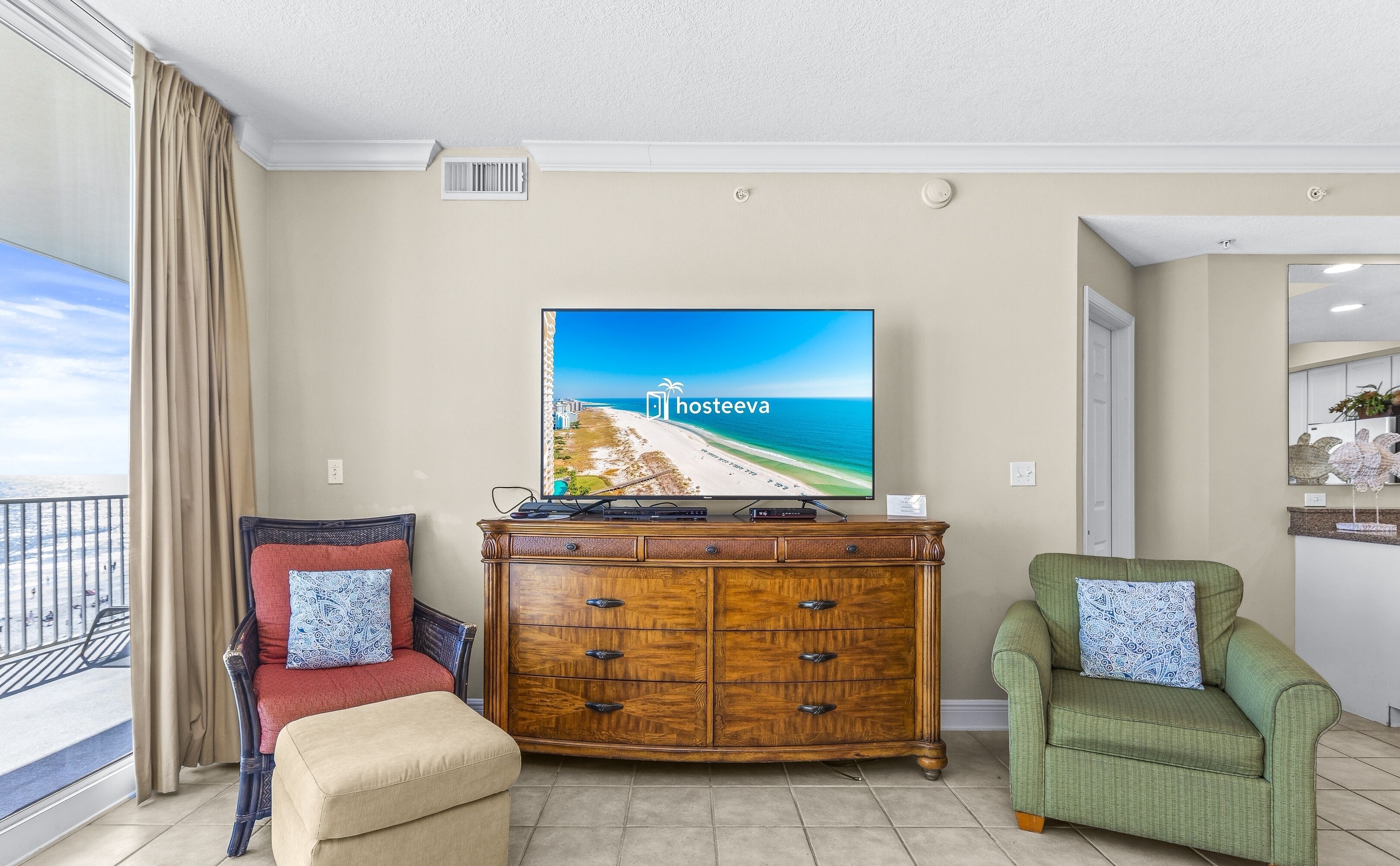 A living room with a large flat-screen TV displaying a beach scene, a wooden cabinet, a red sofa, a green armchair, and a view of the balcony.