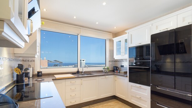 Une cuisine moderne avec des meubles blancs, un réfrigérateur noir et une grande fenêtre donnant sur la mer.