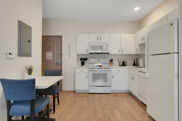 A modern kitchen with white cabinets, a white refrigerator, a stove, and a microwave. There is a dining table with blue chairs and a potted plant on the wall.