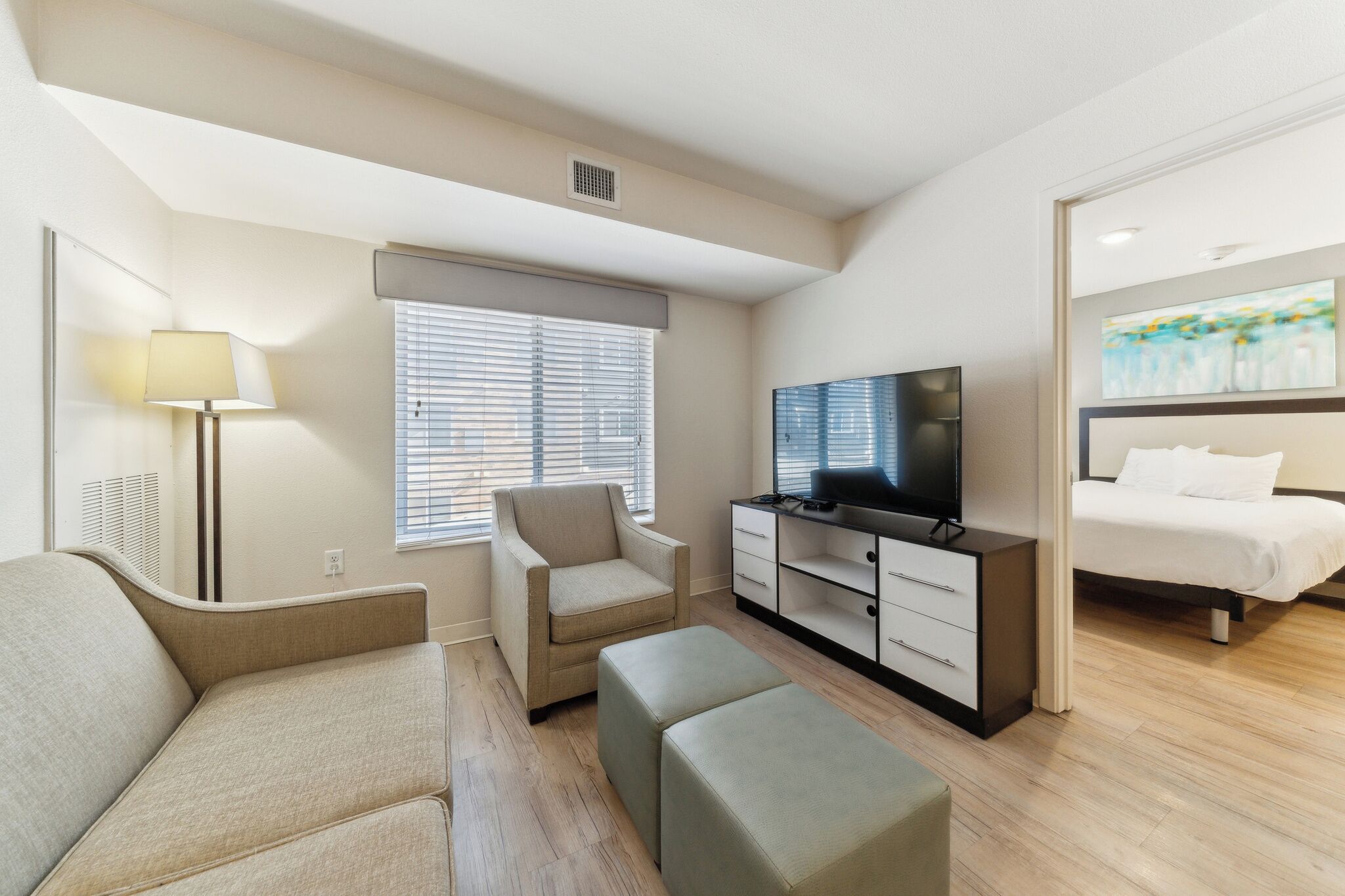 A modern hotel room with a sofa, armchair, ottoman, TV, and a bed.