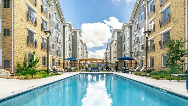 A swimming pool surrounded by multi-story buildings with balconies, outdoor seating, and a covered area.