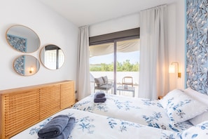Una camera d'albergo con due letti, una testiera in legno, un balcone con tavolo e sedie e vista sul verde.