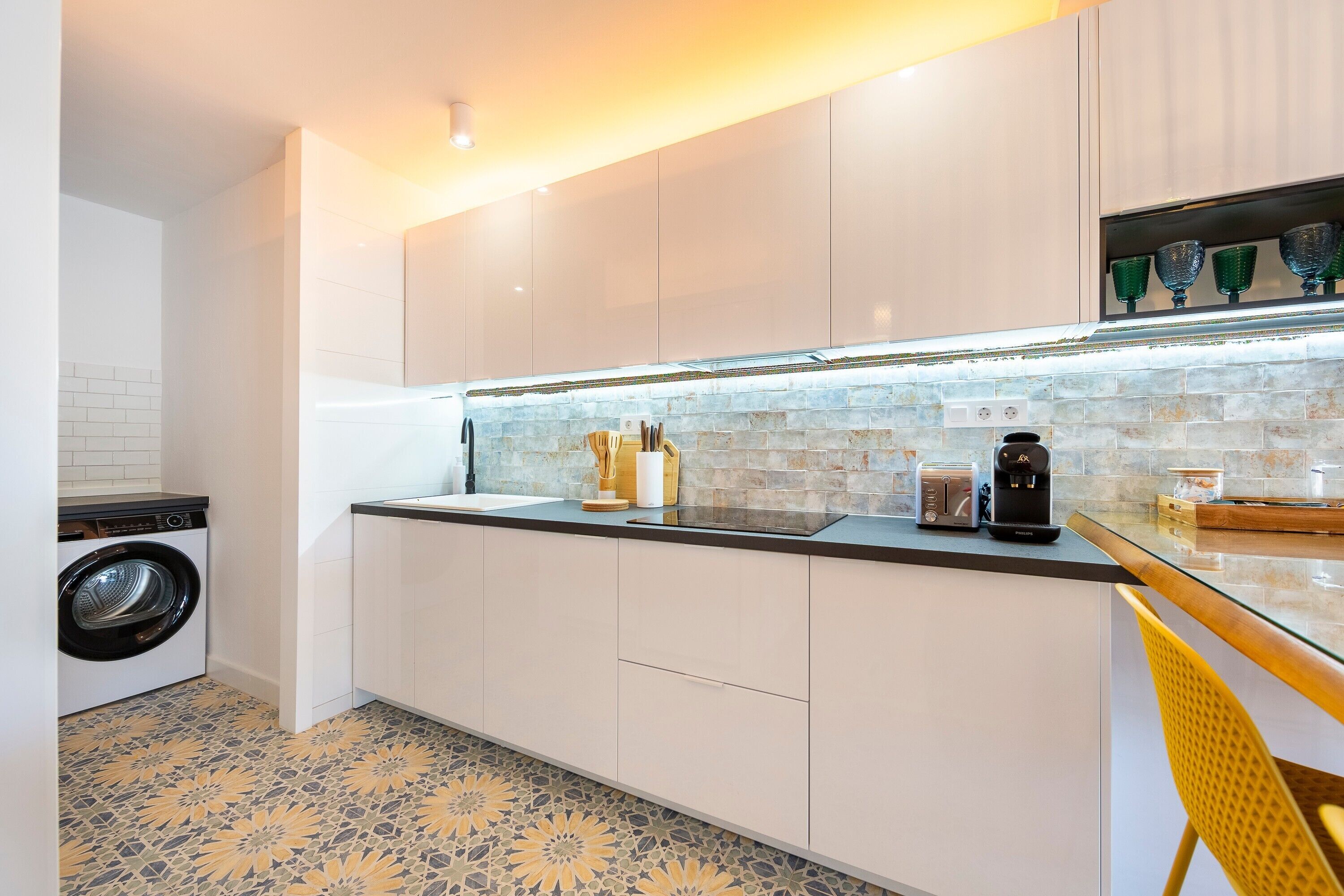 Una cocina moderna con armarios blancos, lavadora integrada, cafetera y una mesa de vidrio.