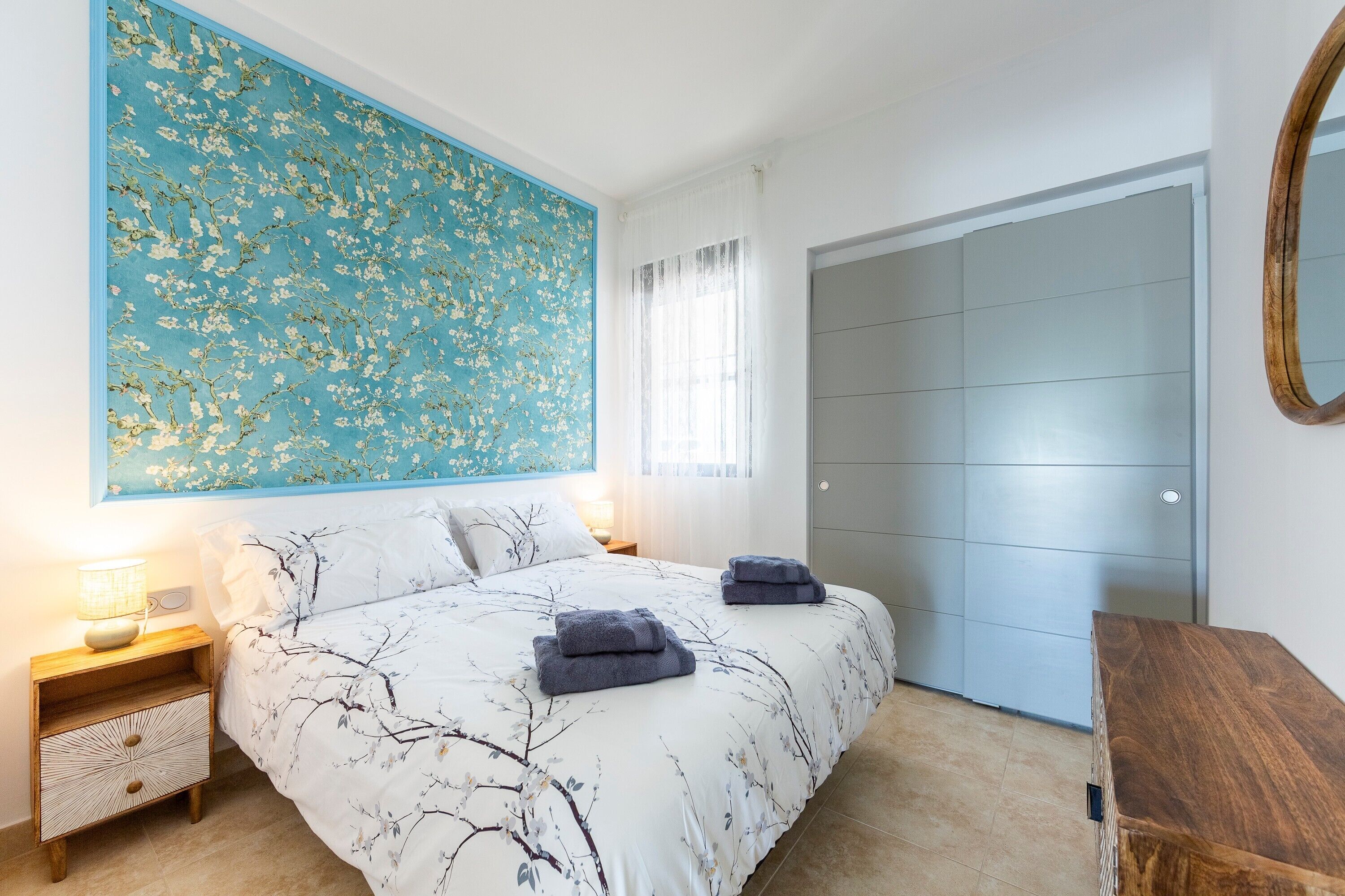 Een slaapkamer met een bed, een nachtkastje, een wandspiegel, een kledingkast en een groot bloemen schilderij.