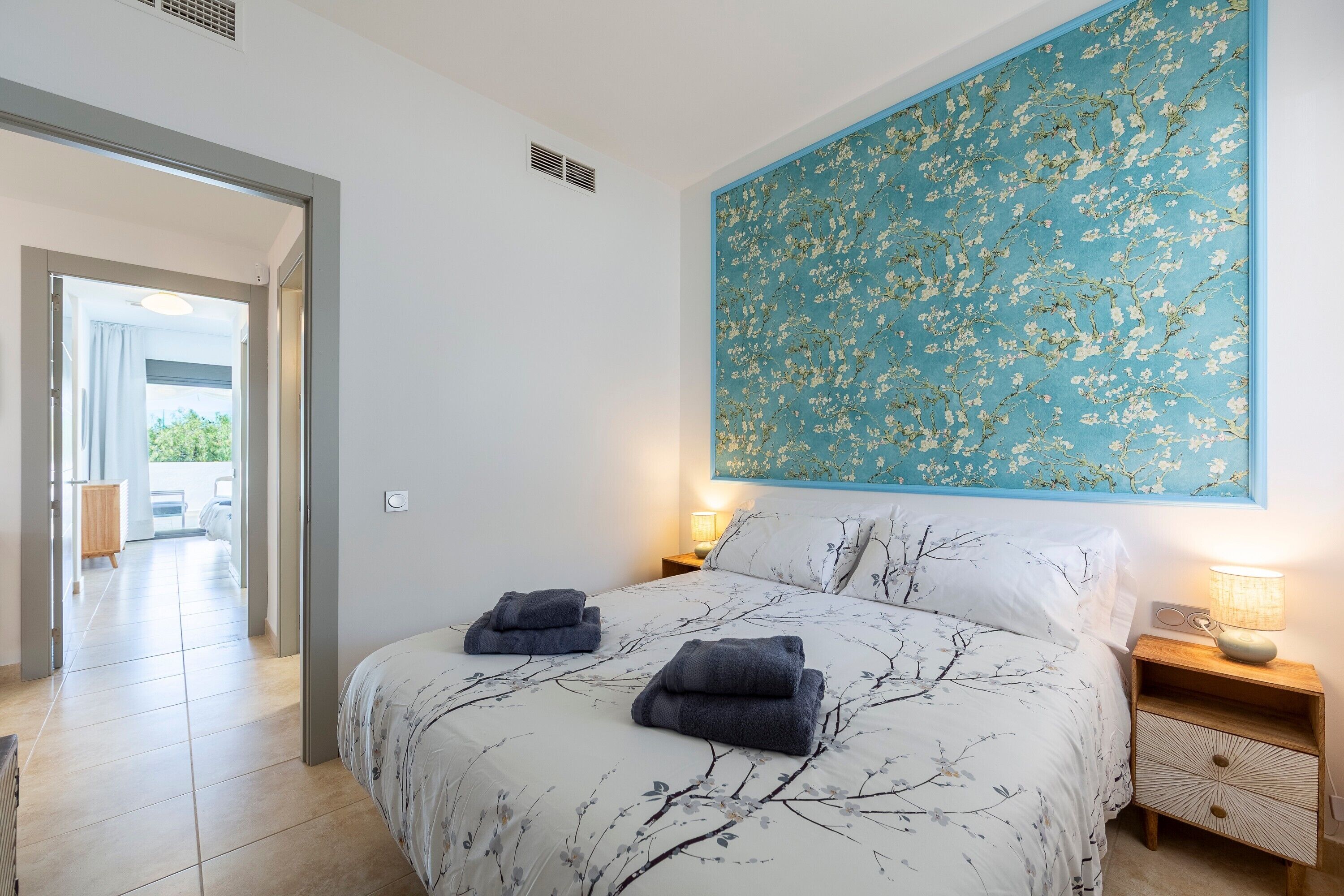 A bedroom with a large bed, two bedside tables, a wall-mounted painting, and a view through an open door.