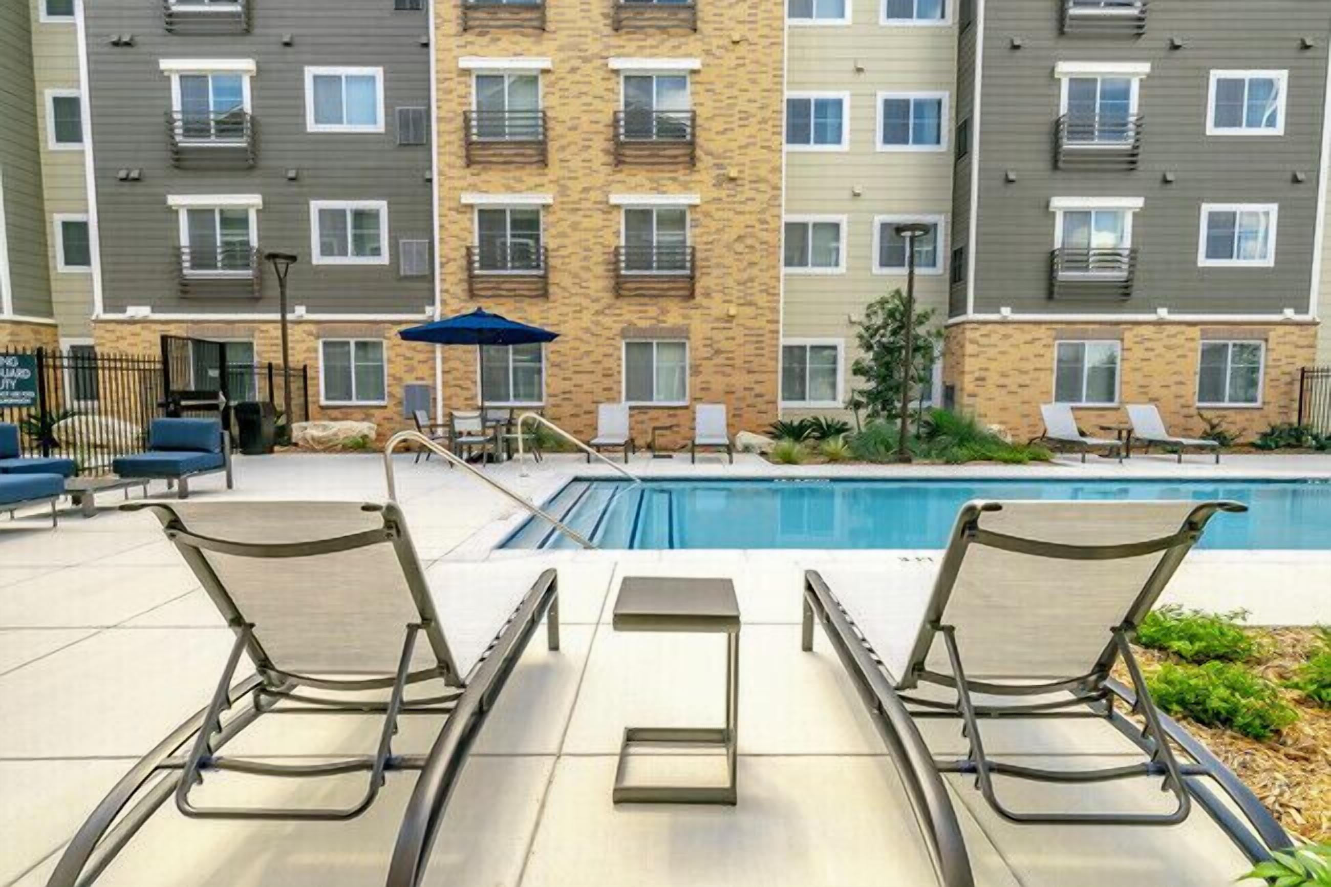 Área de piscina com espreguiçadeiras e uma pequena mesa, cercada por um edifício com várias andares e varandas.