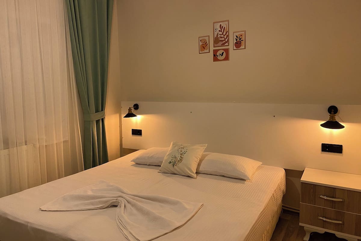 Una cama bien hecha con edredón blanco y una toalla blanca doblada, dos lámparas de noche, una mesita de noche con cajón, y una pared con tres cuadros enmarcados.