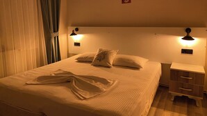 Una cama bien hecha con edredón blanco y almohadas, dos lámparas de noche, una mesita de noche con cajón y una lámpara de pared.