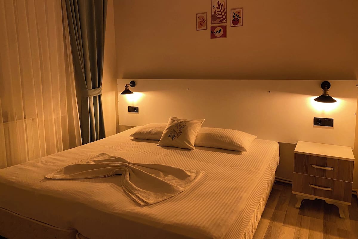 Una cama bien hecha con edredón blanco y almohadas, dos lámparas de noche, una mesita de noche con cajón y una lámpara de pared.