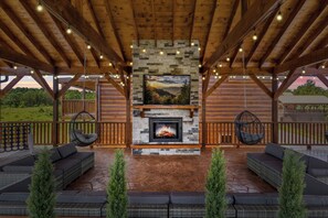 Un espacio de estar al aire libre cubierto con chimenea de piedra, vigas de madera y luces colgantes.
