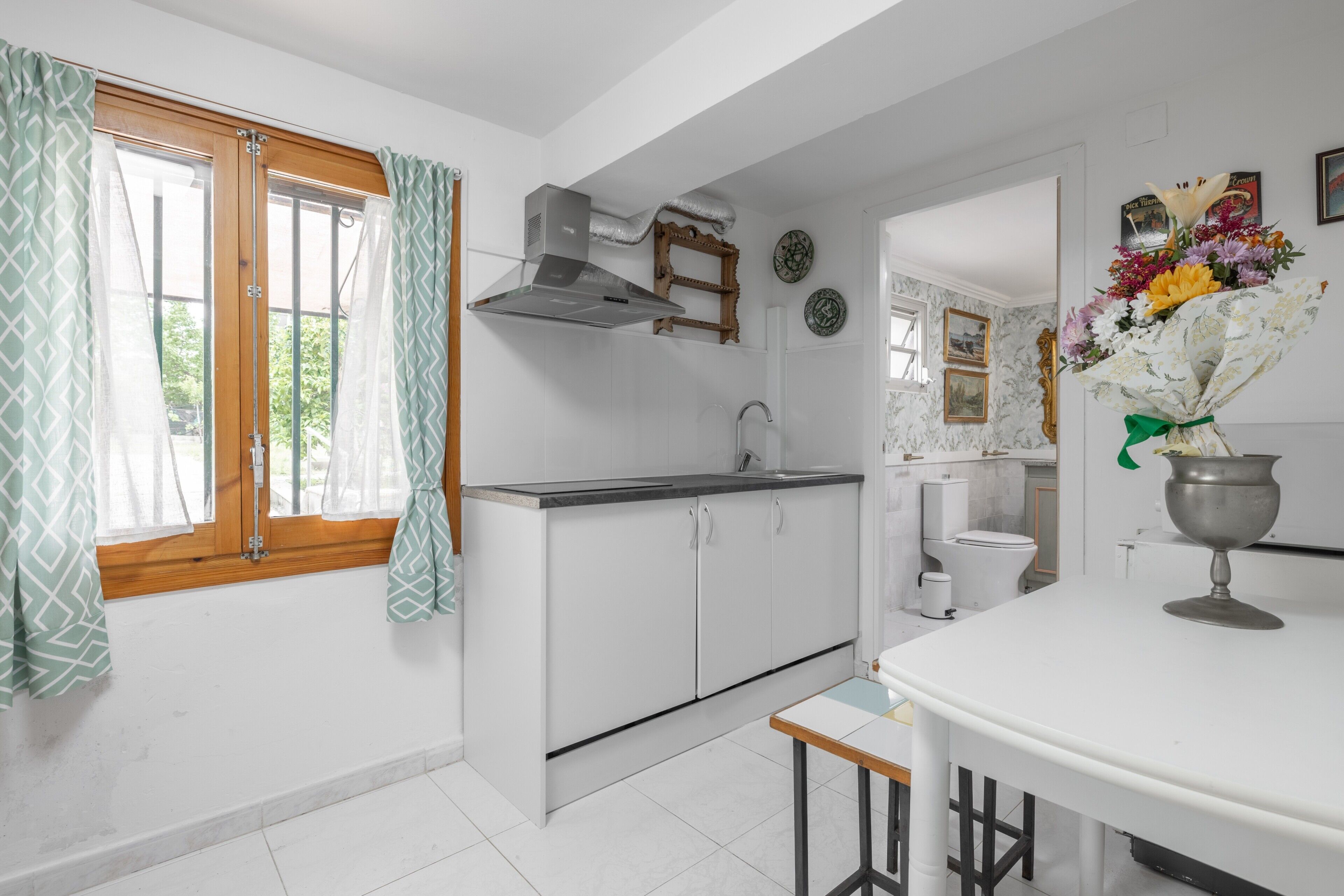 Une cuisine avec des armoires blanches, une hotte de cuisinière en acier inoxydable, une fenêtre ornée de rideaux à motifs et un vase rempli de fleurs posé sur le comptoir.