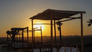 A wooden outdoor seating area with white cushions, a pergola, and palm trees against a sunset backdrop. - Agafay Tenere Luxury Camp (Agafay)