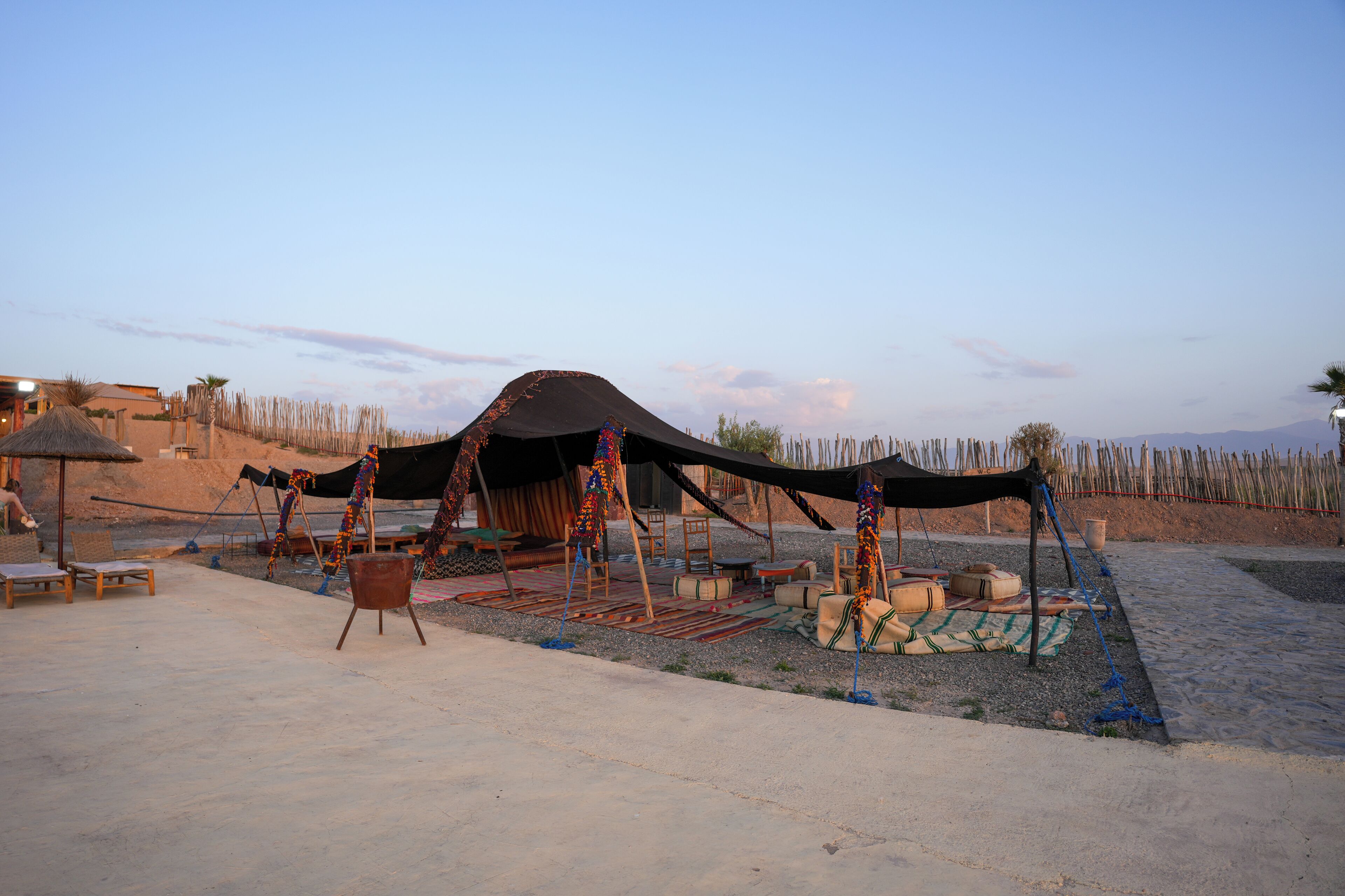 Un campamento en el desierto con una gran carpa negra, sillas de madera y un tapete tejido.