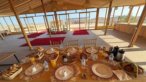 A dining table set for breakfast with various food items and beverages, located in a spacious room with large windows and a view of the desert landscape. - Agafay Tenere Luxury Camp (Agafay)
