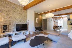 A living room with a stone wall, a flat-screen TV, a sofa, a chair, and a dining area in the background.