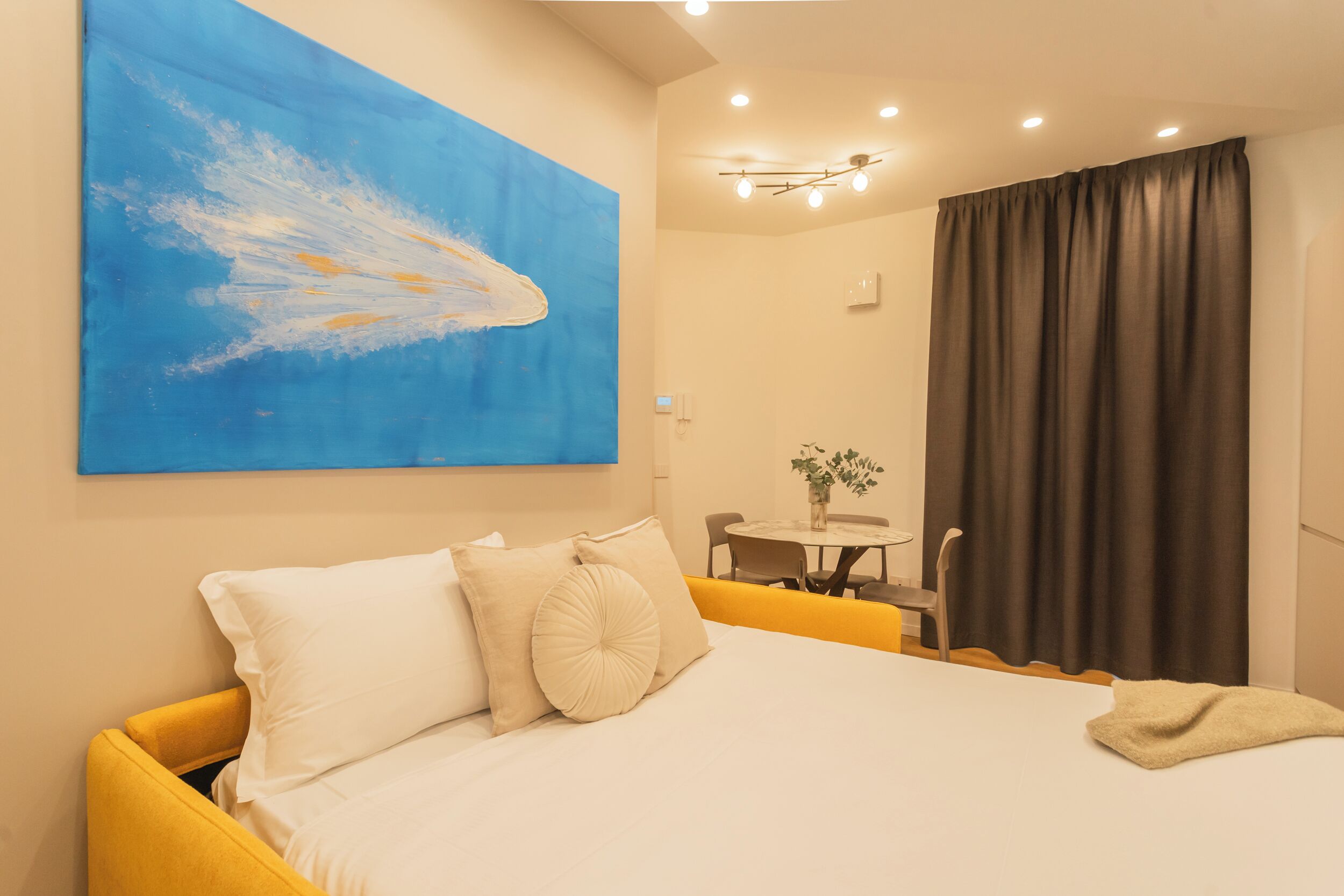 A hotel room with a bed, a painting on the wall, a dining area, and a curtain.