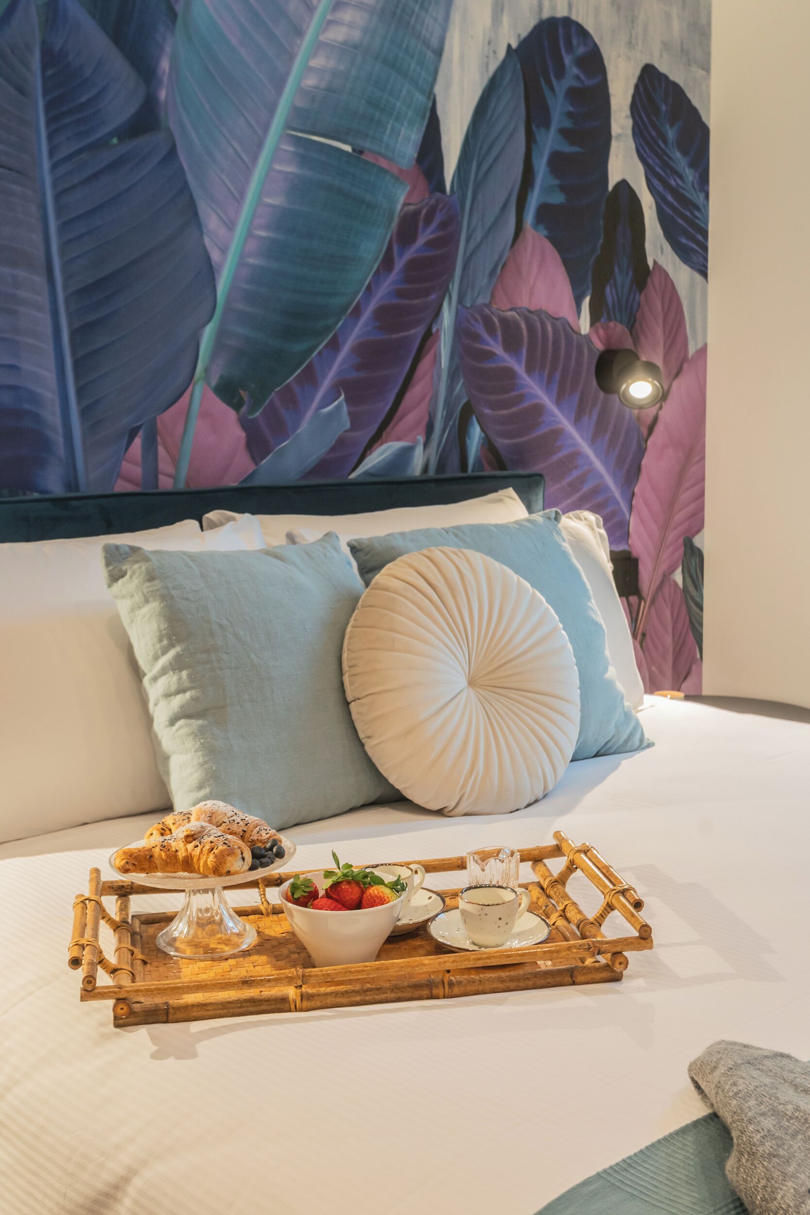 A neatly made bed with a tray of breakfast items, including croissants, strawberries, and a cup of coffee, set against a backdrop of a large, colorful leaf-patterned wall.