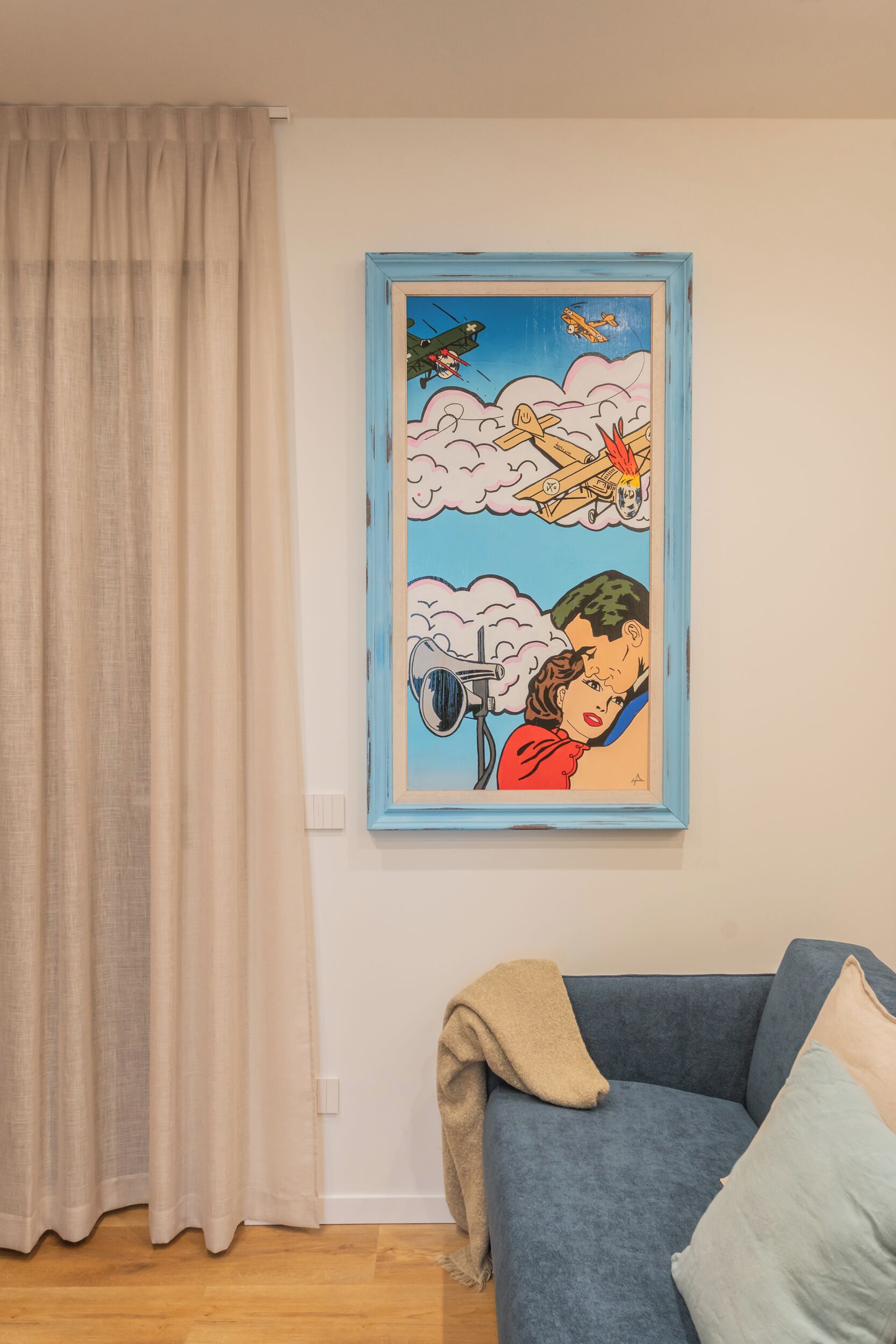 Una sala de estar con un sofá azul, una manta color beige y un cuadro de arte pop en la pared.